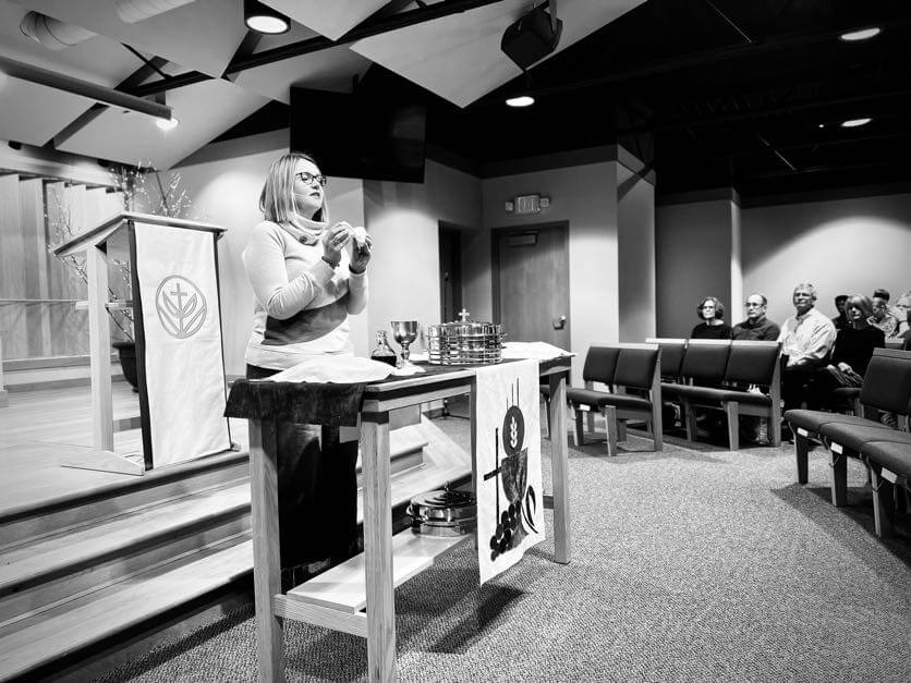 A woman stands behind a communion table in a church, addressing a seated congregation. The scene is in black and white.