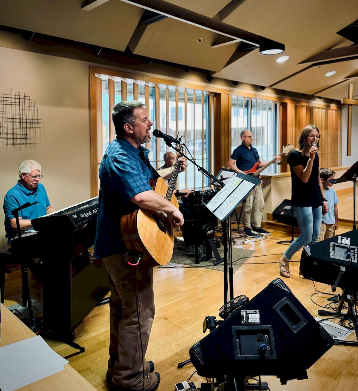 A band performs indoors; a man sings and plays guitar, a woman sings, a pianist plays keyboard, and two others play bass and drums.