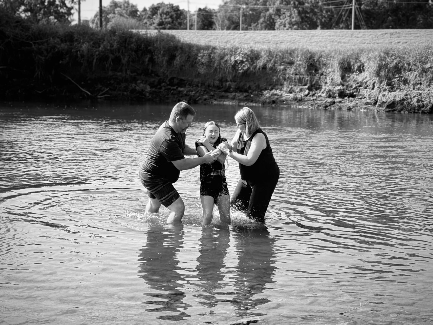 Three people stand together in shallow water, holding hands and smiling, with ripples surrounding them and grassy land in the background.