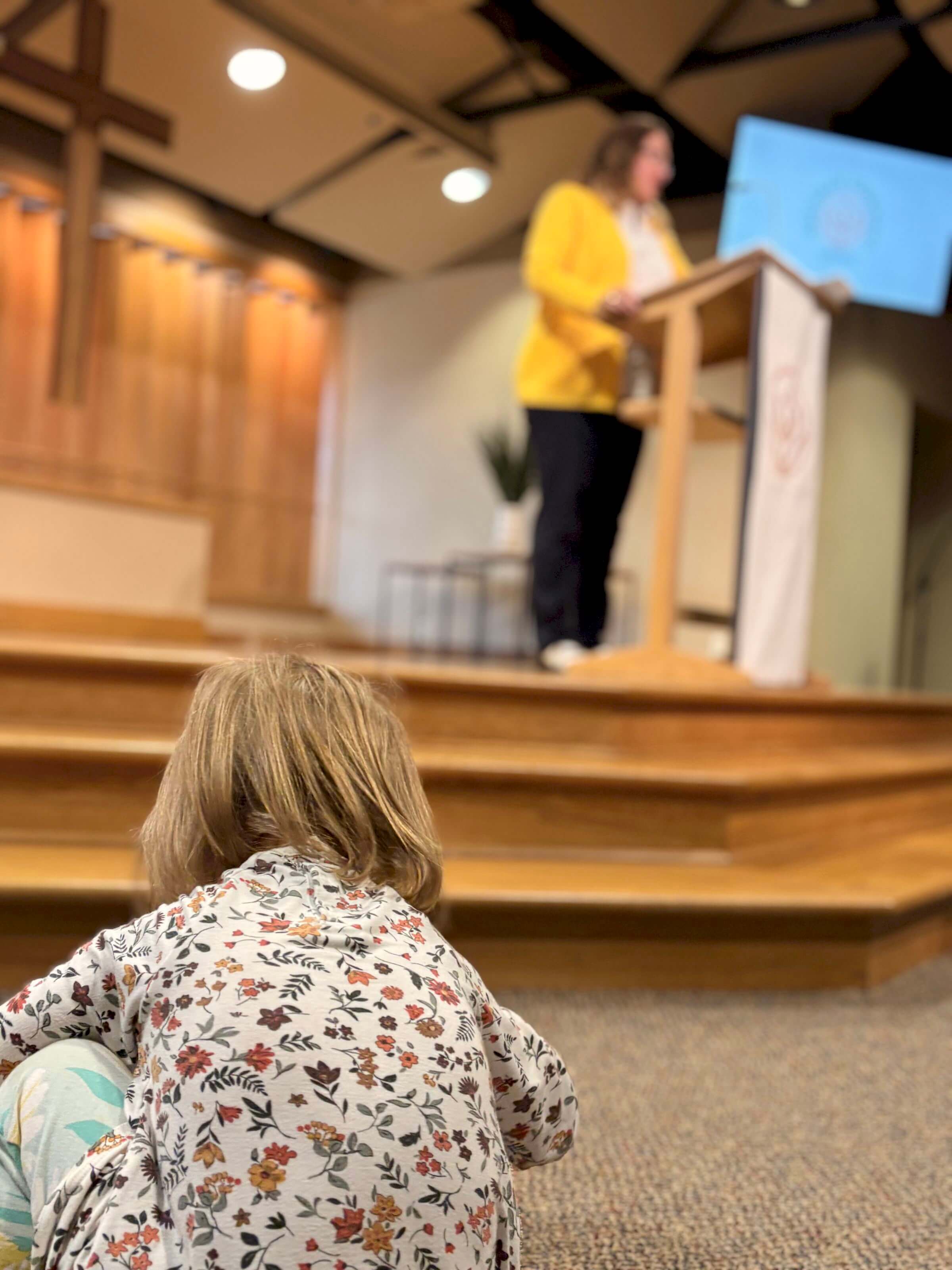 A child sits on the floor, watching a woman speak at a church podium.