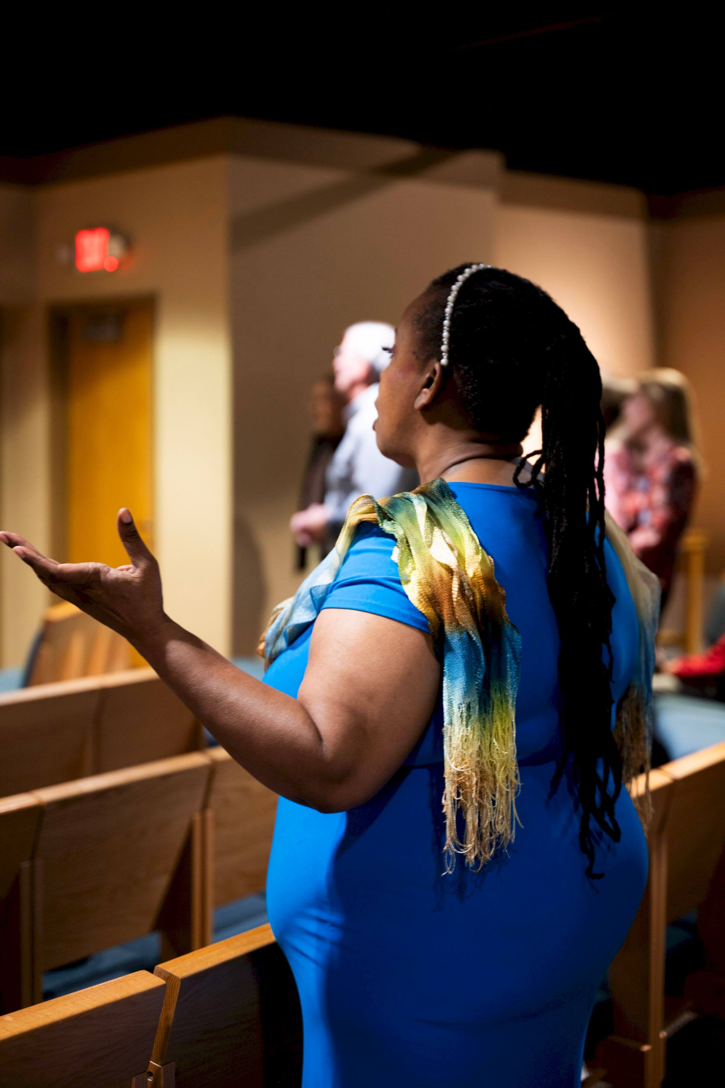 Woman in blue dress with colorful scarf, arms raised, in a church with pews.