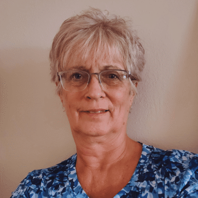Older woman with short gray hair, glasses, and a blue patterned shirt, smiling at the camera.