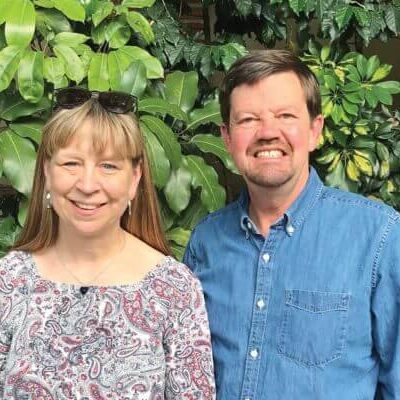 A woman and a man stand side by side outdoors in front of leafy green plants, both smiling at the camera.