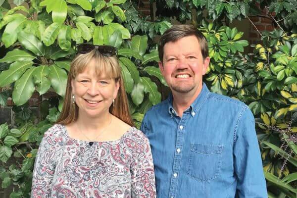 A woman and a man stand side by side outdoors in front of leafy green plants, both smiling at the camera.