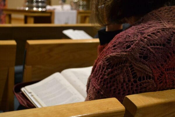 A person wearing a maroon crocheted shawl sits on a wooden bench, reading an open book, possibly a Bible, in what appears to be a church setting.