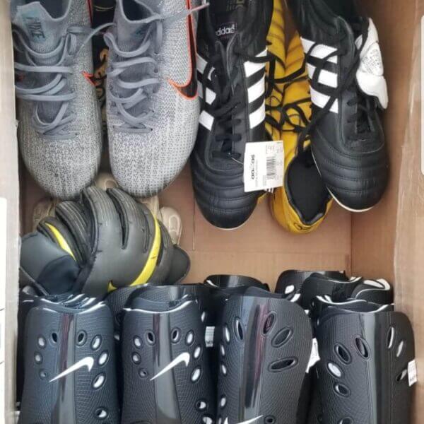 A cardboard box containing three pairs of soccer cleats, two pairs of shin guards, and two pairs of goalkeeper gloves.