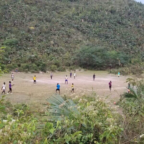 A group of people play soccer on a dirt field surrounded by dense green vegetation and hills.