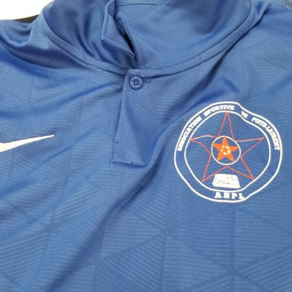 A blue and black collared sports jersey featuring a white Nike logo and a circular patch with a red star, book, and the acronym "ABPL.