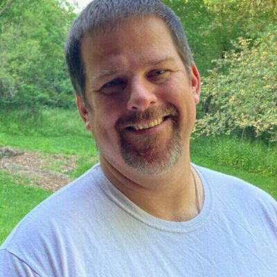 A man with short brown hair and a goatee, wearing a white T-shirt, stands outdoors in front of green trees and grass, smiling at the camera.