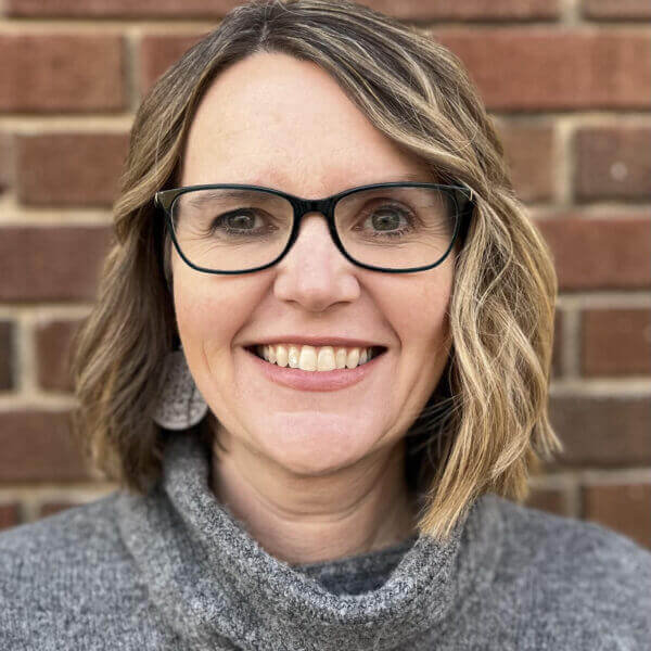 A woman with shoulder-length blonde hair, wearing glasses and a gray sweater, smiles in front of a brick wall.