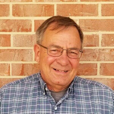 Older man with glasses and a plaid shirt smiles while standing in front of a brick wall.