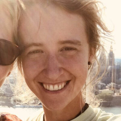 A person with light brown hair smiles at the camera outdoors in bright sunlight, with part of another person visible on the left. A cityscape is blurred in the background.