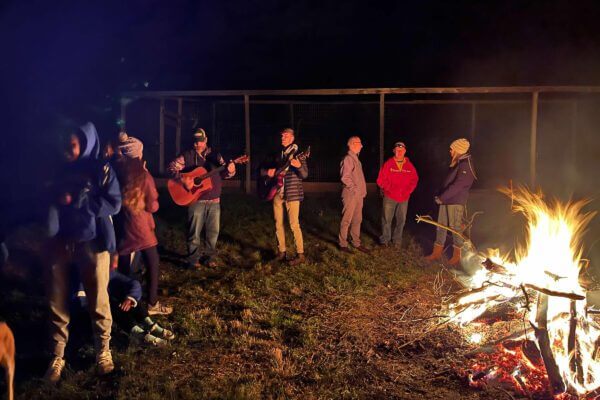 A group of people gathers around a campfire at night; two play guitars while others stand nearby, wearing warm clothing.