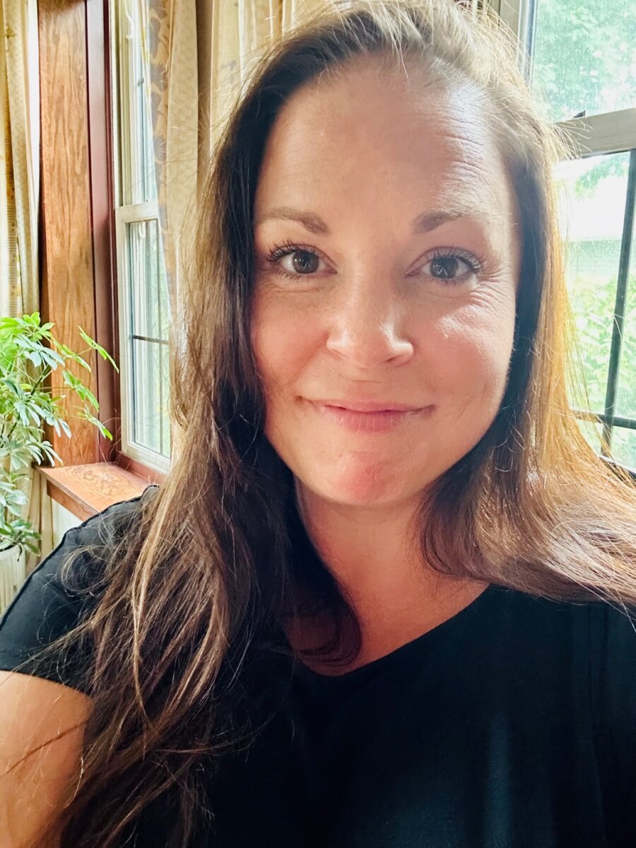 A woman with long brown hair smiles at the camera indoors near a window with sunlight and a green plant visible in the background.