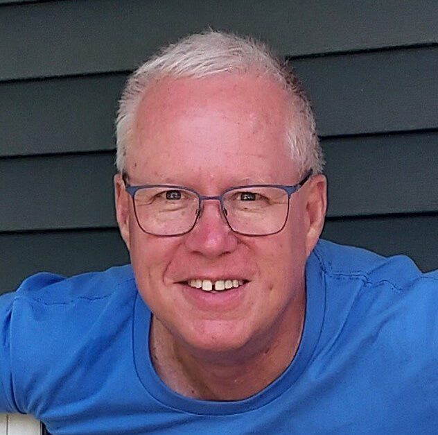 A smiling man with short gray hair and glasses wearing a blue shirt stands in front of a dark green siding background.