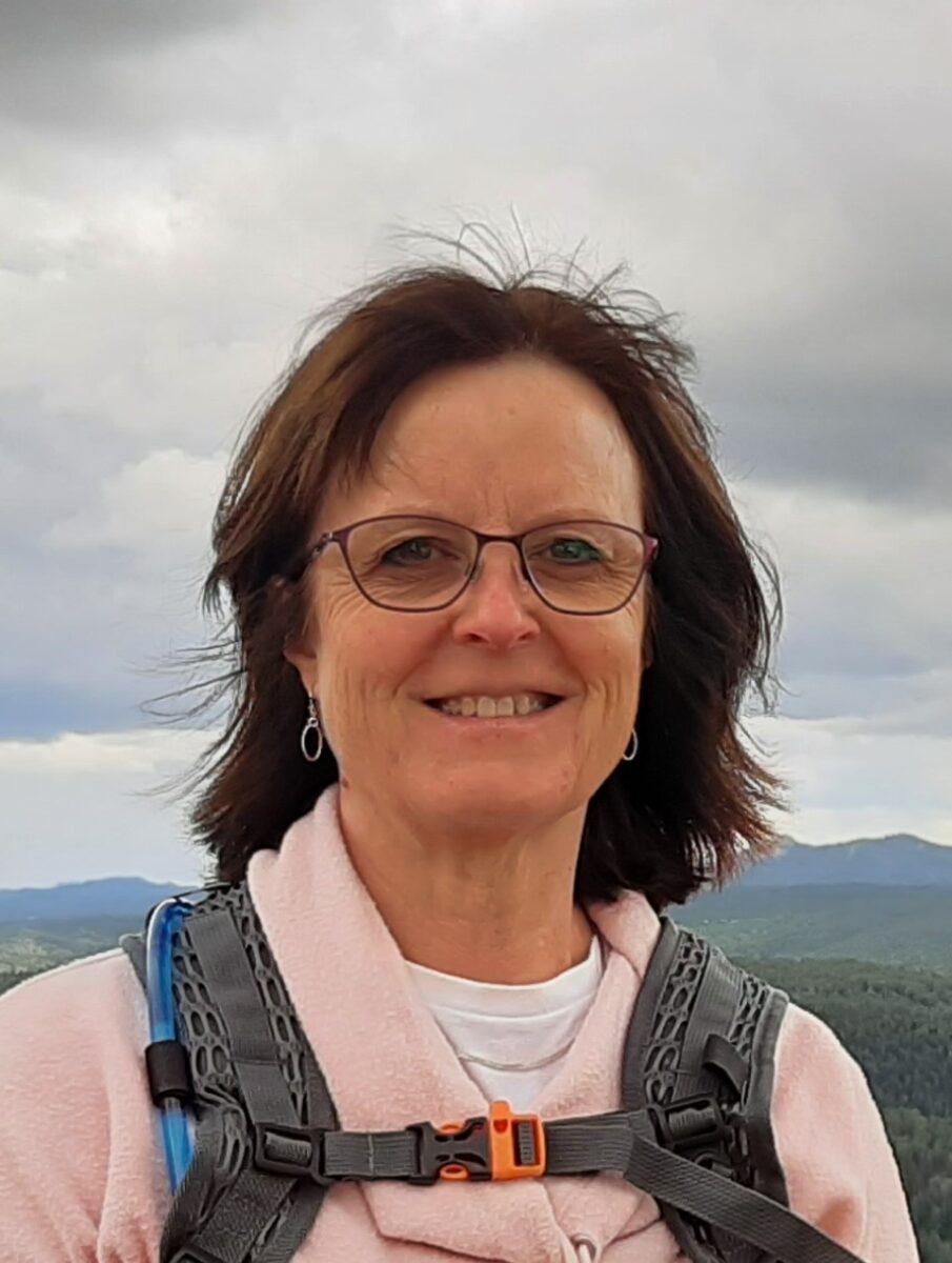 A woman wearing glasses and a light jacket with hiking gear stands outdoors against a cloudy sky and distant mountains.