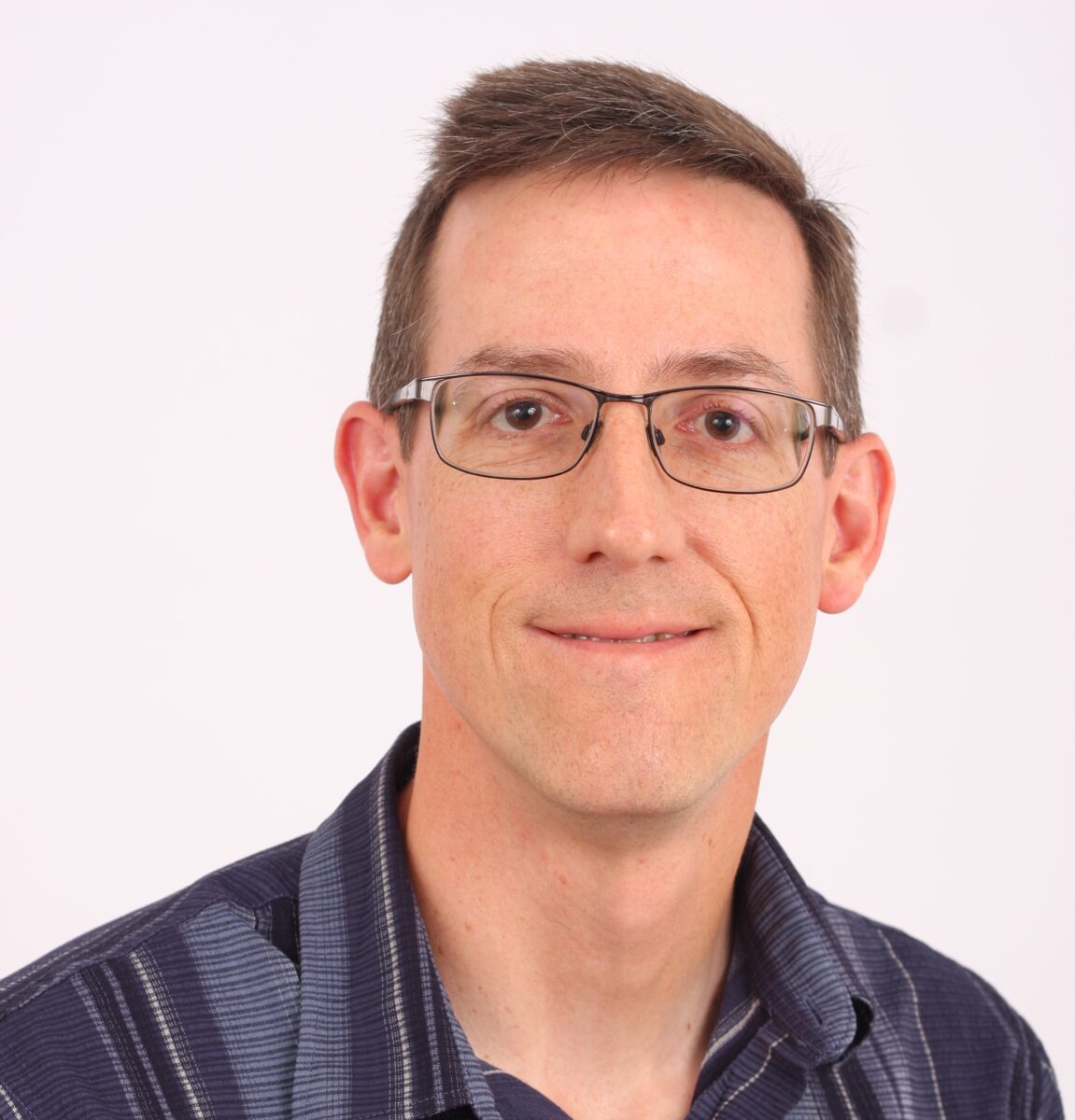 Man with short brown hair and glasses, wearing a dark striped shirt, smiling slightly against a plain white background.