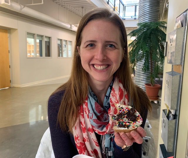 A woman smiles at the camera while holding a sprinkle-covered cookie in an indoor setting with plants and windows in the background.