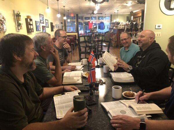Six men sit around a table in a café, reading and discussing books with coffee, pastries, and small Norwegian flags on the table.