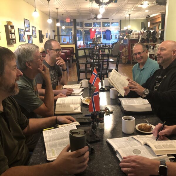 Six men sit around a table in a café, reading and discussing books with coffee, pastries, and small Norwegian flags on the table.