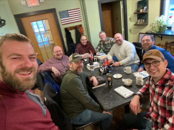 Eight men sit around a black table in a casual restaurant, smiling at the camera. Beverages and food trays are on the table; an American flag hangs on the wall behind them.