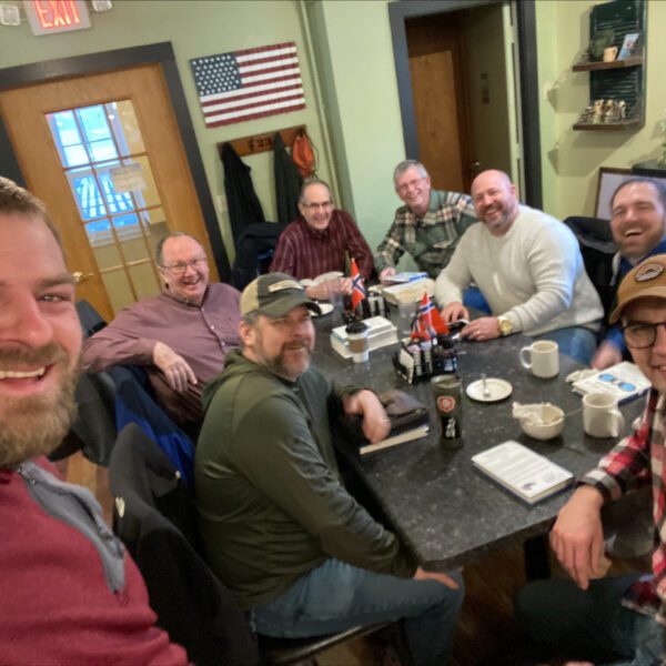 Eight men sit around a black table in a casual restaurant, smiling at the camera. Beverages and food trays are on the table; an American flag hangs on the wall behind them.