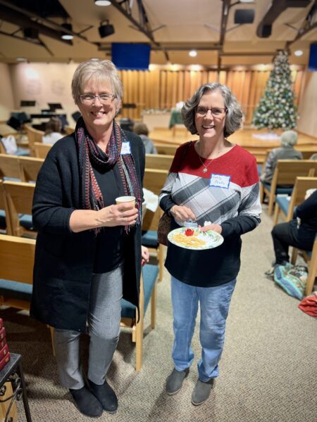 Two women stand smiling in a church hall, one holding a small cup and the other a plate of food, with a decorated Christmas tree visible in the background.