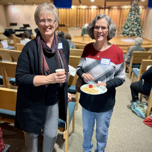 Two women stand smiling in a church hall, one holding a small cup and the other a plate of food, with a decorated Christmas tree visible in the background.