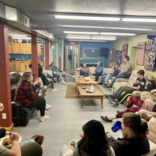 A group of people sit in a circle on couches and chairs in a basement room, talking and eating snacks, with bulletin boards and a kitchen area in the background.
