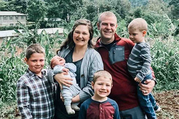 A family of six, including two adults and four children, poses outdoors in front of greenery and buildings on a cloudy day.