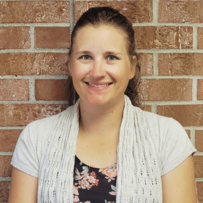 A woman with light brown hair pulled back, wearing a light gray cardigan over a floral top, stands in front of a red brick wall and smiles at the camera.