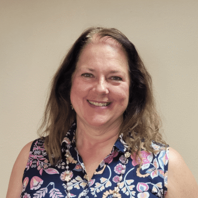 Smiling woman with long brown hair wearing a floral sleeveless top standing against a beige background.