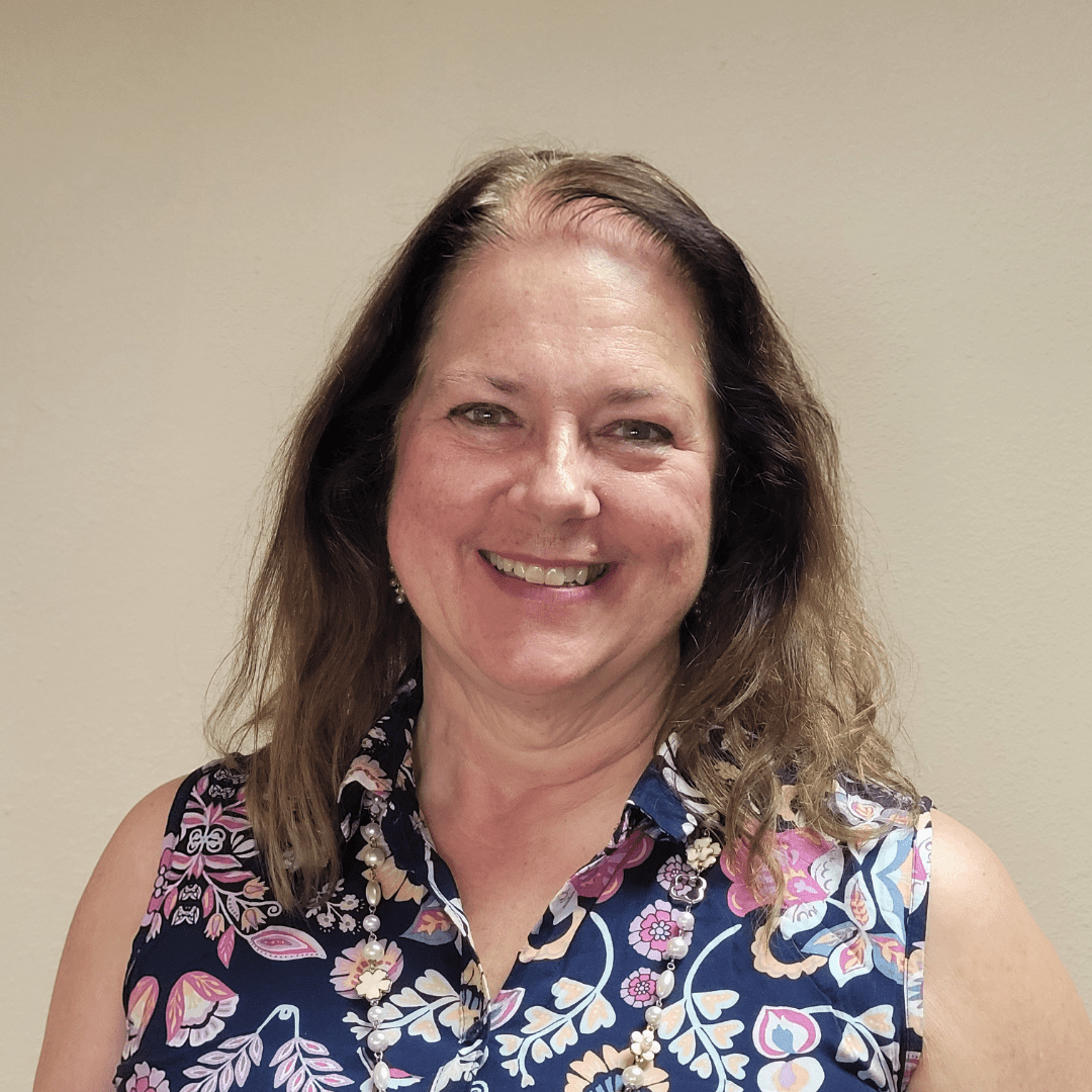 Smiling woman with long brown hair wearing a floral sleeveless top standing against a beige background.