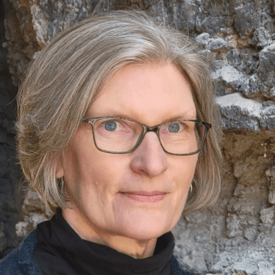 A woman with short gray hair and glasses stands in front of a rocky background, looking at the camera.
