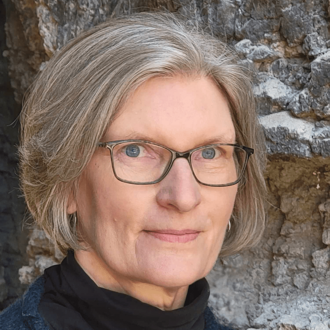 A woman with short gray hair and glasses stands in front of a rocky background, looking at the camera.