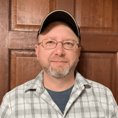 Man with glasses and a cap wearing a plaid shirt, standing in front of a wooden door, smiling slightly.