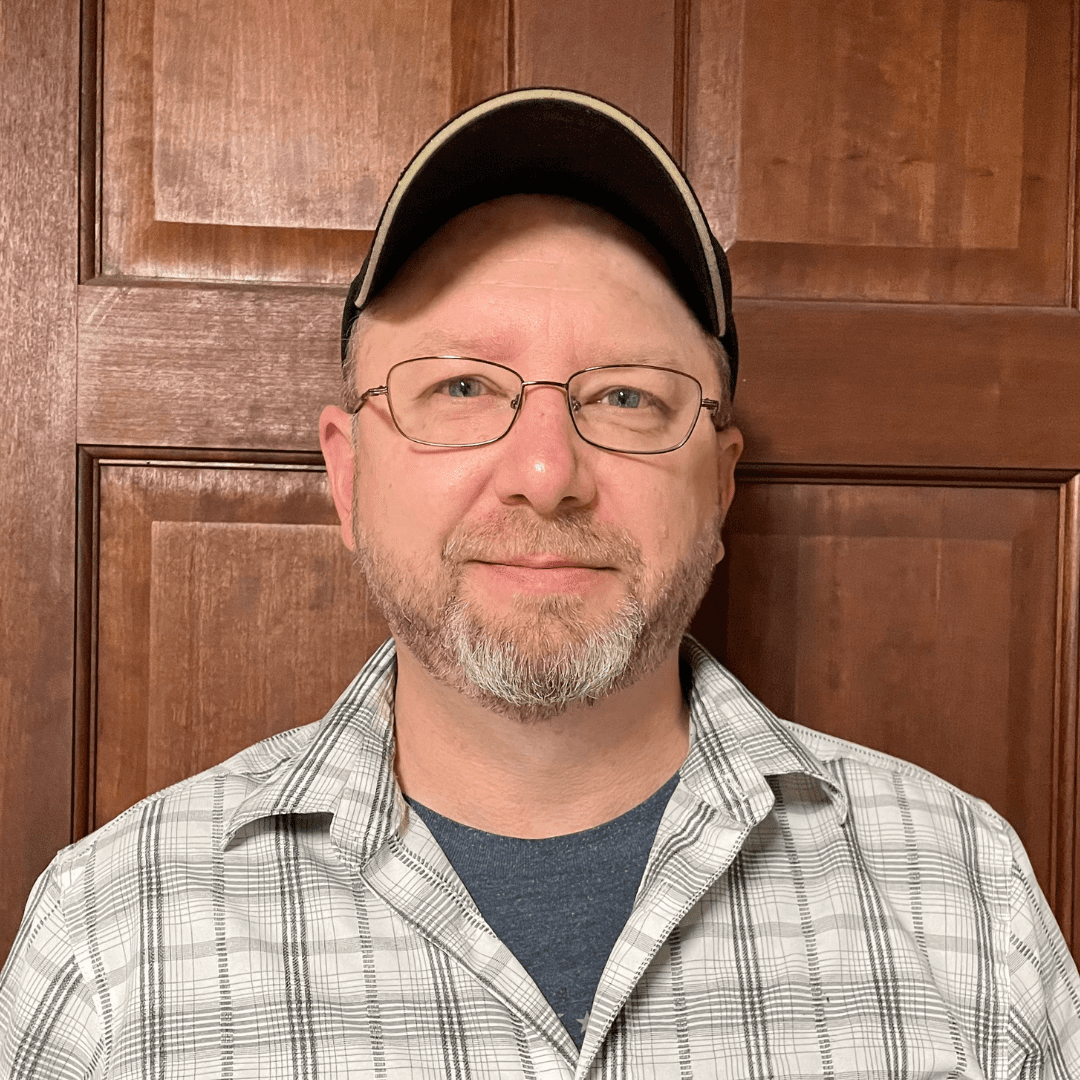 Man with glasses and a cap wearing a plaid shirt, standing in front of a wooden door, smiling slightly.