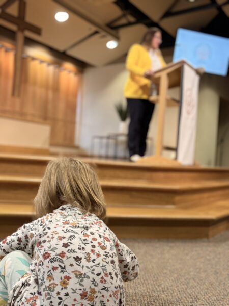 A child sits on the floor as a woman speaks at a podium in a church sanctuary.