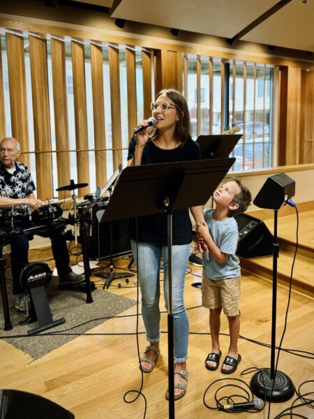 Woman singing into a microphone while a young boy holds her hand and looks up at her; drummer in background.