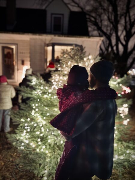 A man holds a child by a lit Christmas tree outside at night, with people and a house in the background.