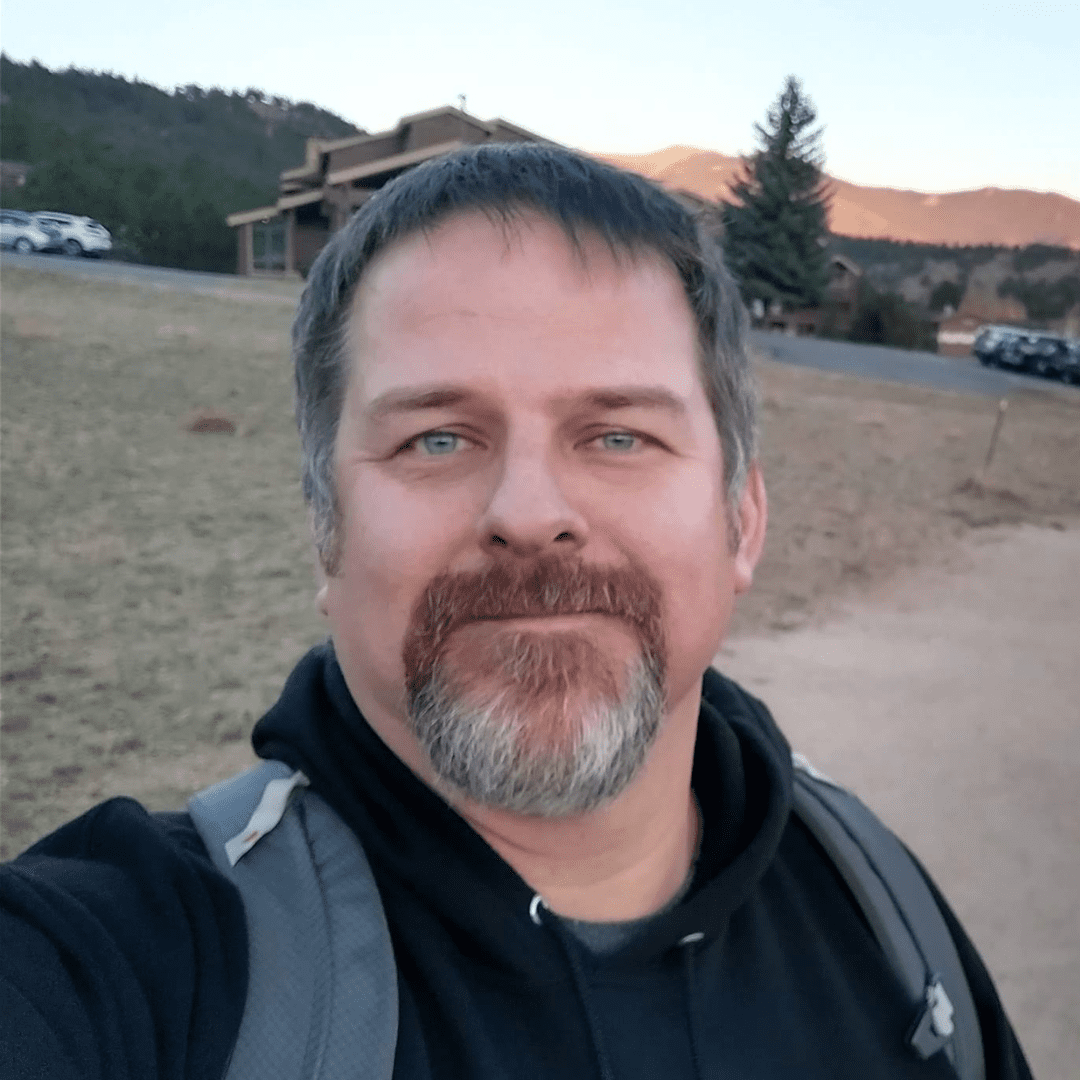 JJ smiles outdoors with mountains and trees in the background.
