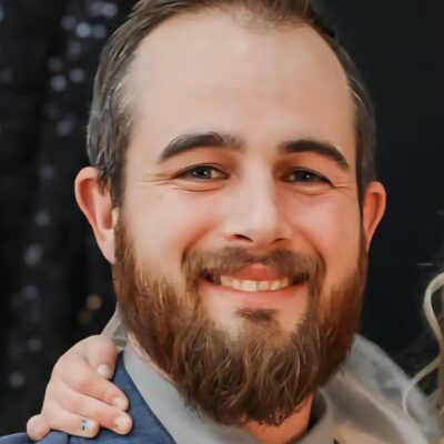 A man with a beard and suit smiling at the camera, with a hand resting on his shoulder.