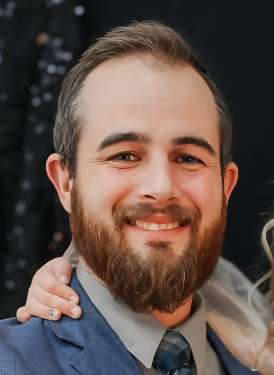 A man with a beard and suit smiling at the camera, with a hand resting on his shoulder.