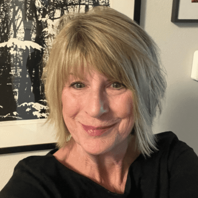 Smiling woman with short blonde hair and a black top, standing in front of framed artwork.