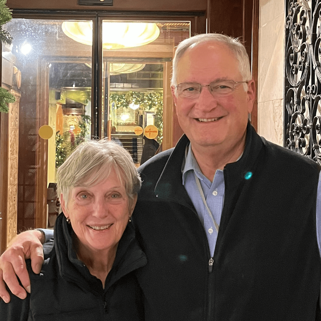Smiling older couple poses together indoors, arm in arm, with festive lights in the background.