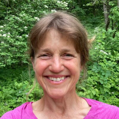 Smiling woman in a pink shirt taking a selfie outdoors with green foliage in the background.