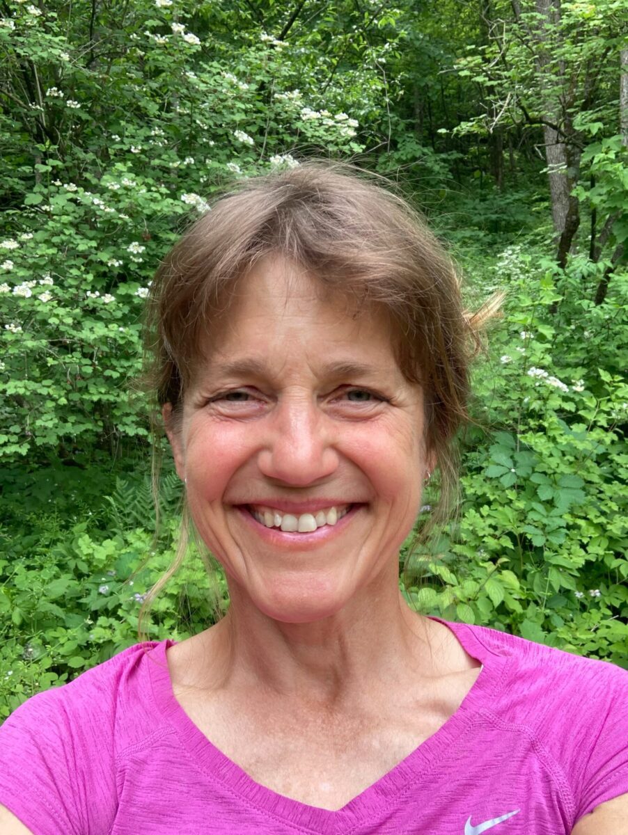 Smiling woman in a pink shirt taking a selfie outdoors with green foliage in the background.