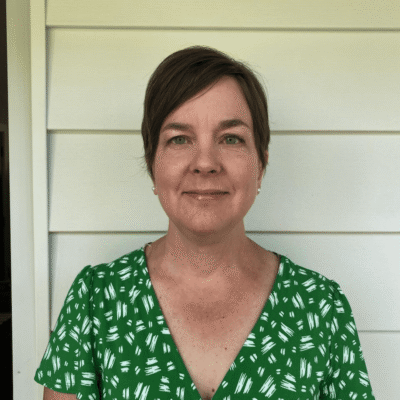 Woman with short brown hair wearing a green patterned dress, standing in front of white siding.