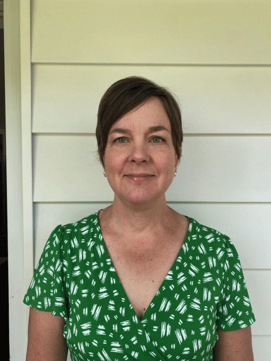 Woman with short brown hair wearing a green patterned dress, standing in front of white siding.