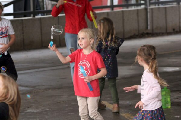 Children play with bubbles outdoors, one girl in a red shirt holds a bubble wand.